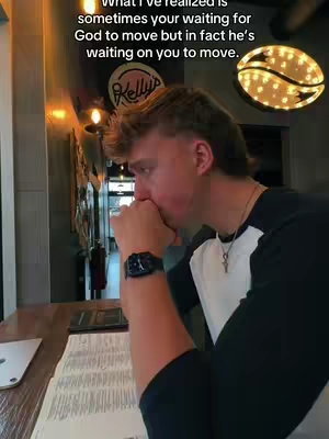 A man with short brown hair wearing a black and white shirt is sitting at a table. He's in partial profile, leaning forward with his hands near his face. The background shows a restaurant interior—a dark wooden table, a menu, and some wall decor. A sign saying 'Kelly's' is visible, and a coffee-bean-shaped light hangs in the background. The overall scene has a warm, somewhat moody lighting.