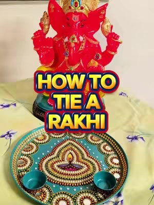 The cover image features a close-up shot of a red, ornate Ganesh statue centered at the top, with the text "HOW TO TIE A RAKHI" overlaid in a colorful, stylized font. Beneath the statue, there's a decorative teal plate with a central design and two small bowls. The background includes a floral patterned fabric.