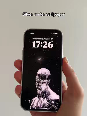 A person's hand holding a smartphone with a silver-colored figure wallpaper against a white background. The time is 17:26.