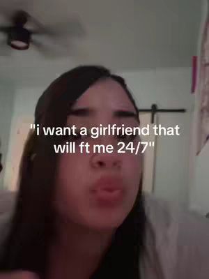 A selfie-style video cover image featuring a young woman centered in the frame. She has dark hair and is looking towards the camera. The background is blurred, suggesting an indoor setting, with visible ceiling fan blades. An overlay text states, "i want a girlfriend that will ft me 24/7". The overall composition is simple, with the focus on the creator and the text.