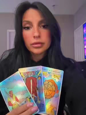 A woman with long dark hair holds up three tarot cards. The cards are brightly colored. The woman is looking at the camera.