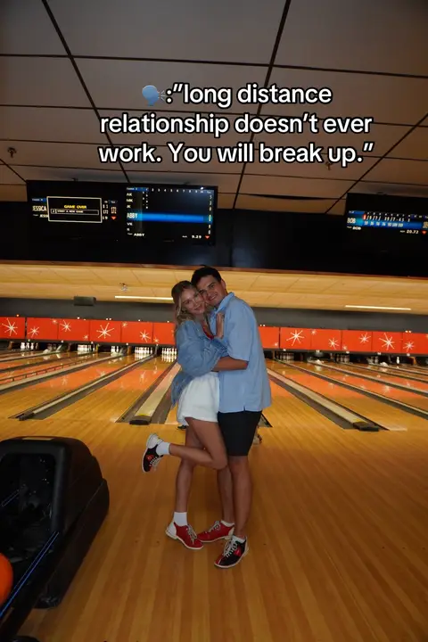 A full shot captures a couple embracing joyfully at a bowling alley. The woman, in a white skirt, stands on one leg leaning into the man. The man, dressed in a blue button-down shirt and black shorts, embraces her tightly. Behind them are bowling lanes. Text is superimposed at the top, indicating a setting.