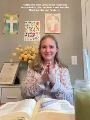 A woman is seated in front of an open book, hands clasped in a prayer position. The background includes artwork on the wall, flowers, and a glass of green liquid, suggesting a morning routine associated with faith and reflection.