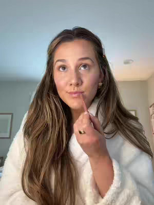 A young woman with long brown hair is applying lip liner in front of a mirror, wearing a white robe. The background includes a light-colored wall and soft warm lighting.