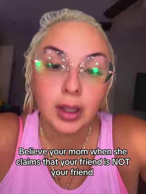 The cover image is a close-up shot of a woman with blonde hair and light skin. She wears large, clear-framed glasses with decorative green accents and a pink top. Text overlayed at the bottom reads, 'Believe your mom when she claims that your friend is NOT your friend.' The background is softly lit, and the overall composition is focused on the woman's face, with her expression being central.