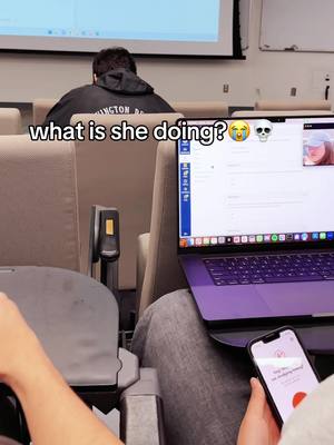 The image shows a classroom setting. A person is seen from the back in a black hoodie, their head obscured. In front of them, a laptop displays a screen with an interface, next to another laptop. The image is lit with ambient light.