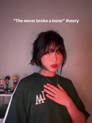 A young woman with dark hair and bangs is centered in the frame. She wears a green shirt with white stripes on the shoulders and a logo that reads 'MAI'. A small shelf with figurines is partially visible in the background, alongside a warm-toned wall. The text overlayed on the top reads, "The never broke a bone theory".
