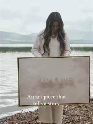 A woman with long dark hair stands outdoors, holding up a framed artwork. The art piece, depicting a light-colored, blurred scene reminiscent of "The Last Supper," is held in front of her. The setting is a serene waterside environment, with calm water and hints of a shoreline in the background.