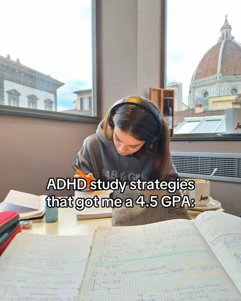 The image is an indoor shot, likely a cafe, showing a young woman sitting at a table, focused on studying. She's wearing headphones and writing in a notebook. Several open books, a pencil case, a cup of coffee, and a building facade are visible on the table and in the background respectively. The composition emphasizes her concentration and study environment.