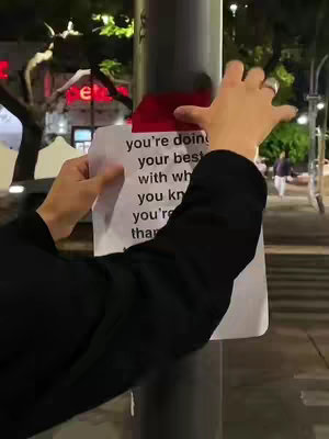 A person holding a white paper with black text, attached to a dark gray pole at night. The background shows blurry city lights and buildings.