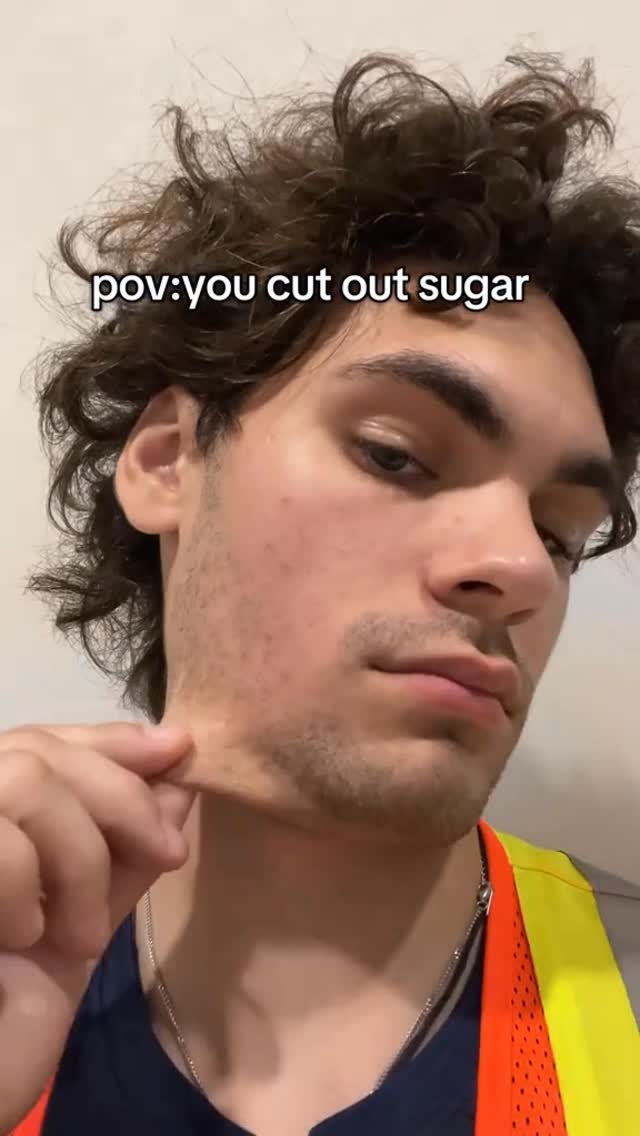 A selfie-style video shows a fair-skinned man holding the skin of his neck. He has wavy, dark hair and is wearing a navy blue top and a safety vest. The text overlay reads: pov: you cut out sugar.