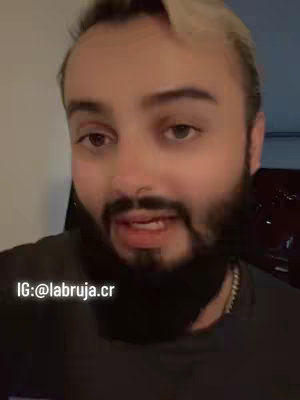 A man with a dark beard and short, styled blonde hair looks directly at the camera and speaks. The background is blurred, with a faint outline of a room visible. Overlayed text at the bottom reads IG: @labruja.cr.