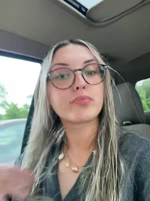 A young woman with long blonde hair and glasses sits in the driver's seat of a car, looking towards the camera. She wears a necklace and a striped shirt.