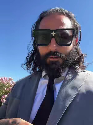 A man wearing large black sunglasses, a suit and a tie, poses against a bright blue sky and some pink flowers.