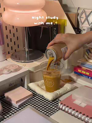 The cover image shows a well-lit desk scene with a person pouring a beverage from a can into a clear plastic cup. The cup is placed on a small, white, square cushion. The desk setting has a lamp with a spherical shade, a stack of books, and a pink notebook. The background includes various stationery items, creating a cozy and organized study environment.