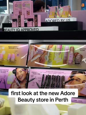 The image is a view inside an Adore Beauty store. The foreground displays product displays with a variety of skincare, makeup, and haircare products. The background features retail displays with lighting.