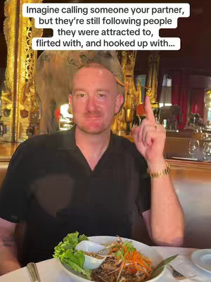 A man, likely in a restaurant, points upwards while looking at the camera. The background shows decorative woodwork and lighting. In front of him is a plate of food.