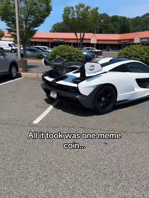 A white McLaren Senna sits parked in a parking lot. The car is angled towards the camera, showcasing its rear and side profile. The image is sunny and bright with a glimpse of a storefront and parked cars in the background.