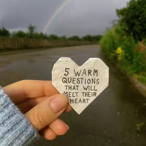 A hand holds a white, heart-shaped piece of paper with handwritten text against a backdrop of an overcast sky and a hint of a rainbow. The hand is holding the heart in front of an out-of-focus scene of a road or path lined with trees, suggesting a natural, possibly outdoor setting.