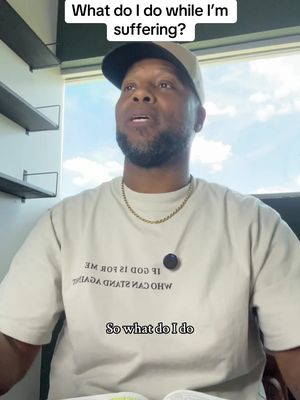 The cover image shows a medium shot of a Black man wearing a black cap, gold necklace, and a cream-colored t-shirt with text. He is indoors, likely in an office, with a window and shelving visible in the background. He is looking upward with a contemplative expression, and overlaid on the top is a white text that says: 'What do I do while I'm suffering?' with another line of text overlaid that says: 'So what do I do'. There is a small black object.
