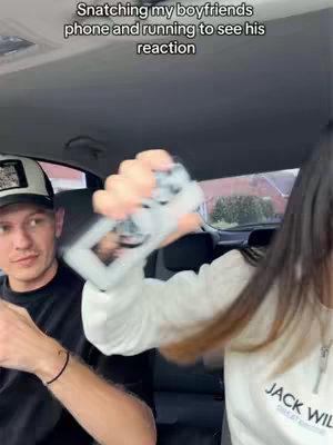 The image shows a woman reaching towards the camera with one arm, grasping a smartphone as if she's about to snatch it. A man, seated in the driver's seat of a car, looks at her with a slight smile, arms at his side. Text overlays the image, indicating a scene of playful interaction.