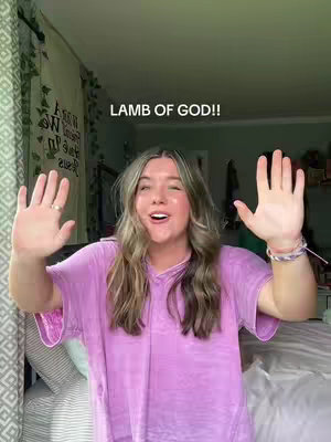A young woman with long blonde hair stands with her arms outstretched, palms facing the camera. She's smiling and appears animated. She wears a pink t-shirt in a bedroom setting. The text "LAMB OF GOD!!" is written in uppercase above her head.