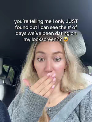 A woman with long blonde hair, wearing a gray sweater is in the driver's seat of a car, possibly a selfie video, she's covering her mouth with her hand in an expression of surprise. Text overlays the image in black font.