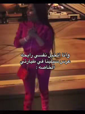 The image shows a woman standing on what appears to be an airport tarmac. She has long dark hair and is wearing a pink athletic outfit. She is looking to the left and appears to be speaking. The background is blurry, showing a plane, airport equipment, and the evening sky. There is superimposed text in Arabic script.