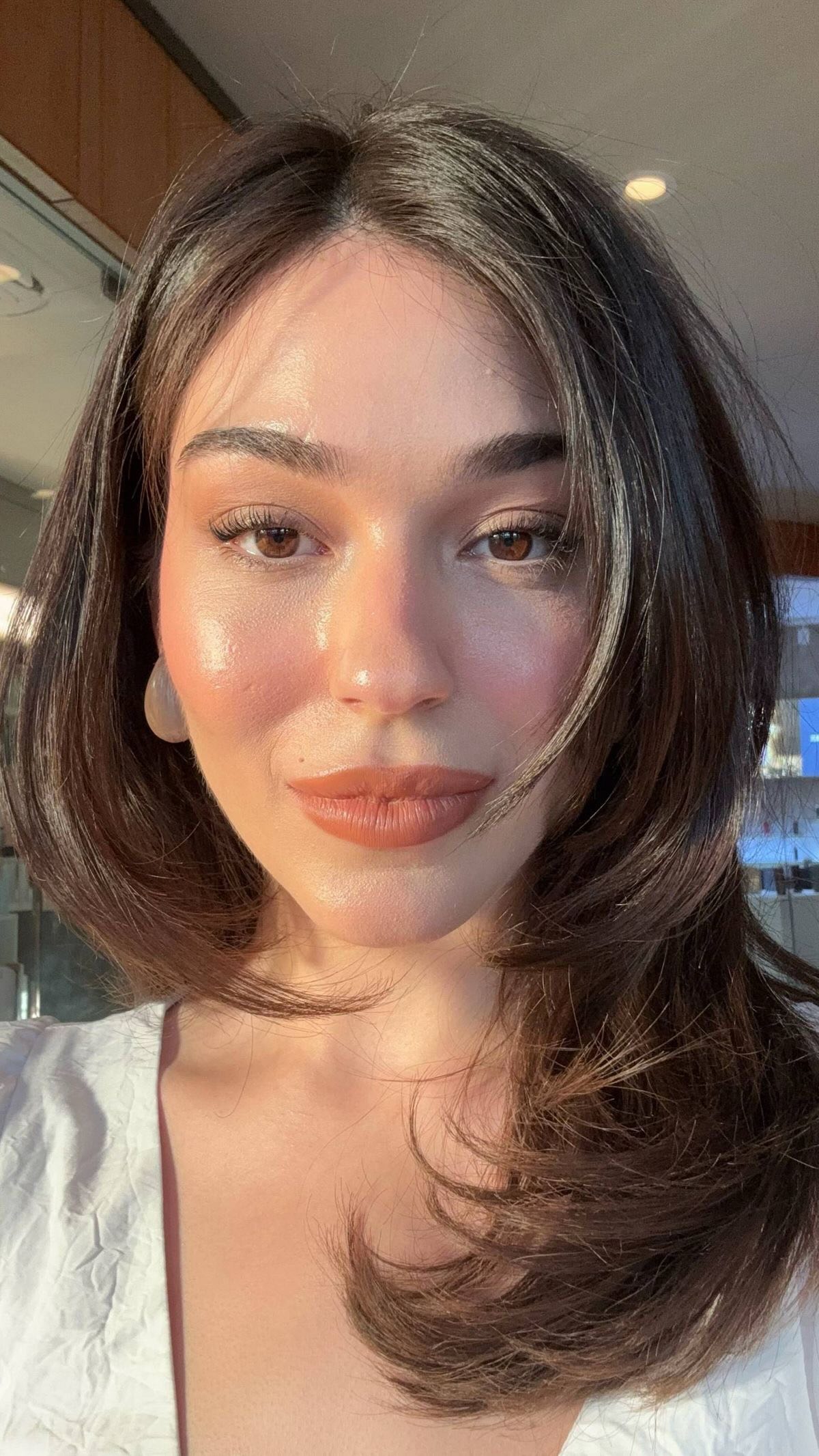 A close-up selfie featuring a woman with dark hair, warm-toned skin, and makeup. She is looking directly at the camera with a soft smile. The image is well-lit, and the background is slightly out of focus.