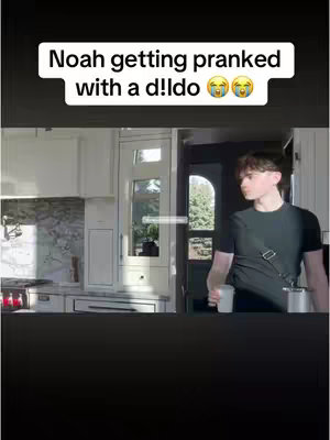 A young man stands in a kitchen, looking slightly to the side. He's holding a white cup or mug and is dressed in a black t-shirt. The kitchen has white cabinets and a light streaming in from a window. A text overlay indicates the video is about a prank.