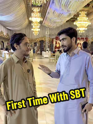 Two men in traditional Pakistani clothing inside a decorated venue, with ornate chandeliers and seating visible in the background. The foreground features the phrase "First Time With SBT" in a yellow outlined font.
