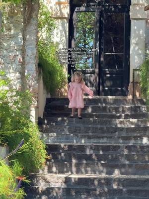 A young girl in a pink dress descends stone steps. Behind her is a glass door and a stone wall with lush greenery. The sunlight creates deep shadows, and the composition has a dreamy, inviting quality.