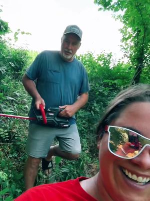 A selfie-style image with two people in a green, natural environment. A woman with sunglasses is in the foreground, smiling widely. The sunglasses reflect a distorted image. Behind her, a man is seen holding a hedge trimmer, appearing to clear a path. The background is filled with dense greenery, suggesting a wooded area or overgrown path.