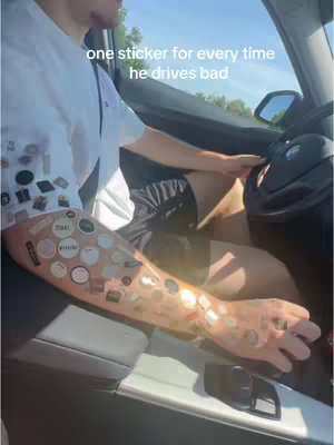 A person is sitting in the driver's seat of a car, arm resting on the door, with stickers covering most of the visible portion of the arm. The scene is inside the car, with the road visible outside.