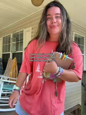 A person with long hair and a red shirt stands outdoors, holding a clipboard covered with colorful clips and stickers. The person smiles looking toward the camera. The background is a wooden exterior.