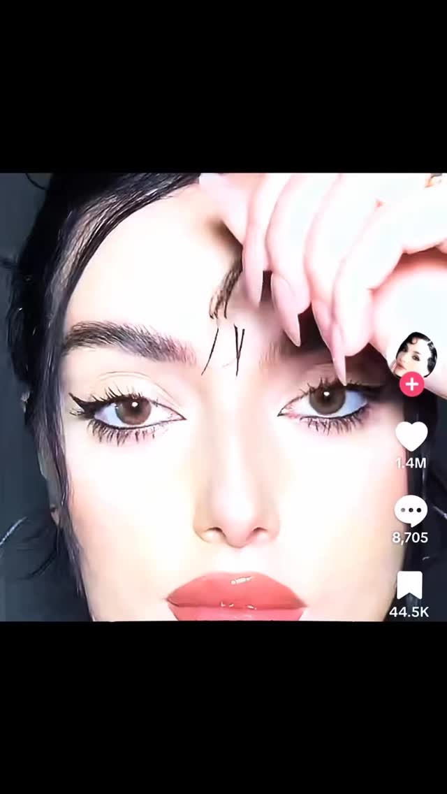 A close-up shot of a woman's face, focusing on her eyes and lips. She has dark hair, and her eyes are heavily lined with black eyeliner, with long, dark eyelashes. A small portion of a hand with manicured nails is visible near her eyebrow with lines drawn on her forehead. The lips are glossy and full. The background is dark, without distractions, giving focus on the woman’s face.