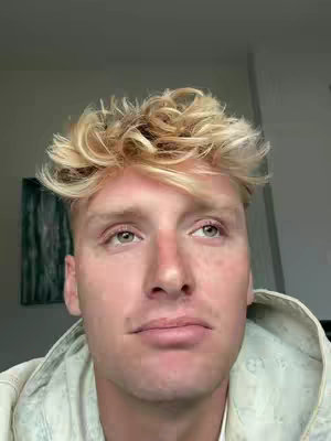 A close-up shot of a fair-skinned man with light blonde, tousled hair. He looks upward and slightly to the side with a pensive expression. He is wearing a light-colored, hooded jacket, and a partly obscured painting hangs in the background.