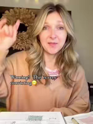 A woman with long blonde hair points upwards with her right index finger. The image is a mid-shot with text overlaid on the bottom left corner. The background is blurred, but includes a decorative wreath.