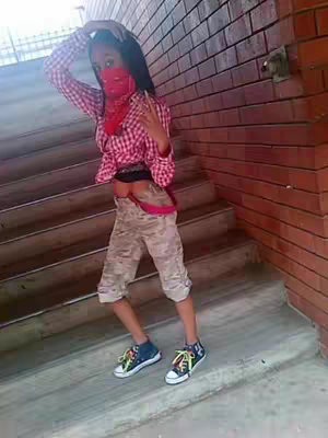 A young person, appearing to be of mixed heritage, poses in a full body shot taken on a concrete staircase. They are wearing a combination of urban and casual clothing: camouflage patterned shorts, a red and white checked button-down shirt cropped at the midriff, and colorful sneakers. A protective face covering in a similar shade conceals the lower part of their face. Their right hand is positioned in a peace sign. The background features both the staircase and a red brick wall.