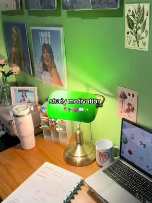 A desk setup with a green lamp, a laptop, a mug, a notebook, and other study-related items. There are also framed pictures and artwork on the wall. The overall color scheme is dominated by green and warm wood tones.
