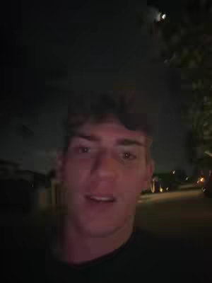 A selfie-style shot of a young man's face, filling most of the frame. His expression is neutral, with his eyes open and looking slightly upwards. The background is dark, suggesting nighttime, and somewhat out of focus, featuring indistinct shapes of buildings and foliage. The lighting is soft, with the man's face being the most illuminated element.