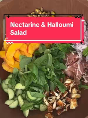 A top-down shot of a wooden bowl filled with a colorful salad. The salad consists of various ingredients arranged in sections: sliced peaches and halved cherry tomatoes on the left, arugula and fresh mint in the center, prosciutto and red onions to the right and pieces of halloumi on the bottom, and pistachios at the top.