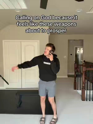 A man in a black hoodie, gray shorts, and sandals is on a phone call, gesturing with his right arm. The background includes white double doors, a neutral-colored wall, and a section of a staircase.