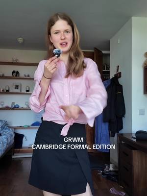 A young woman stands in a room holding a small microphone, appearing to speak. She wears a pink blouse tied at the waist, a black skirt, and a black bracelet. The setting includes a bedroom or living space with shelves and a bed visible in the background, indicating an at-home filming location.