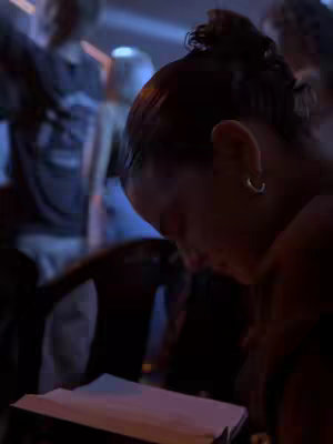 The close-up shot features a person with dark hair held up in a bun or updo, wearing a hoop earring. Only the side profile is visible, and the person is looking down, presumably at a book or paper object held in their lap. The background is blurred, indicating a low depth of field. The lighting appears to be dim, which obscures the face from clear view.