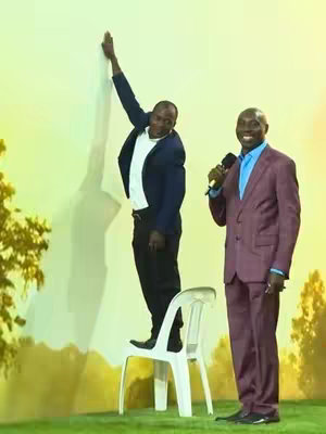 The image shows two men on a stage-like setting. One man is standing on a white chair with his arm raised as if reaching for a wall, and appears to be wearing a dark blue suit with a white shirt. The other man is standing next to him, wearing a burgundy suit with a blue shirt and holding a microphone. The background is a gradient of yellow and green, suggesting a studio or an outdoor setting with a touch of graphic design.