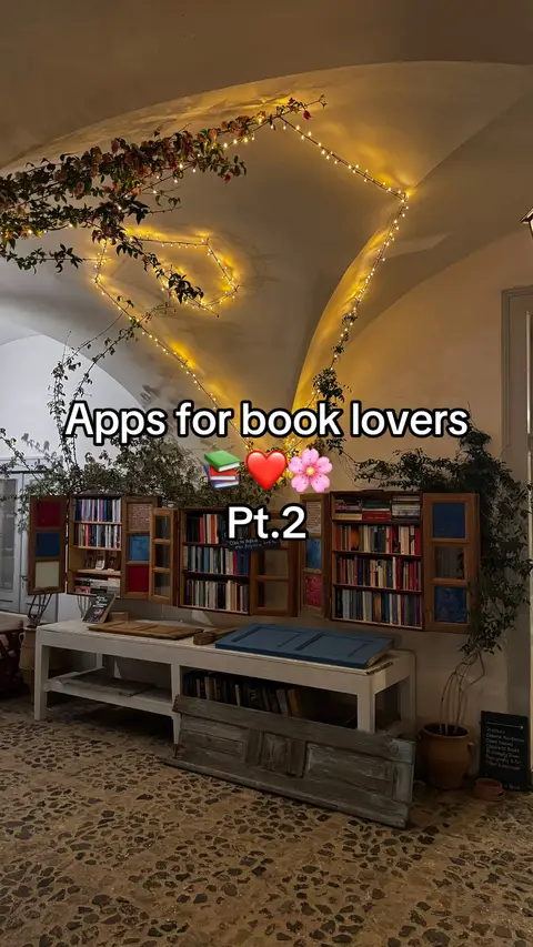 The cover image shows a cozy indoor book nook setting, with a curved, arched ceiling lit by strings of warm, yellow fairy lights. The space has a stone tile floor with visible brick-like patterns. The backdrop features a built-in bookshelf filled with many books, and the text overlay states 'Apps for book lovers' along with the words 'Pt.2' and a collection of emojis. The scene is inviting and warm, suggesting a relaxed environment conducive to reading.