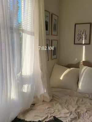 A softly lit bedroom interior with morning sunlight streaming through a sheer white curtain and blinds. The scene includes a bed with white bedding, a window, and framed artwork on the wall. The overall impression is one of a bright, calm morning.