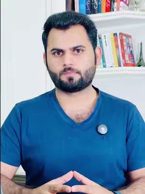 A man with a beard and short dark hair looks directly at the camera, his hands clasped in front of him. The background includes a white bookshelf filled with books.