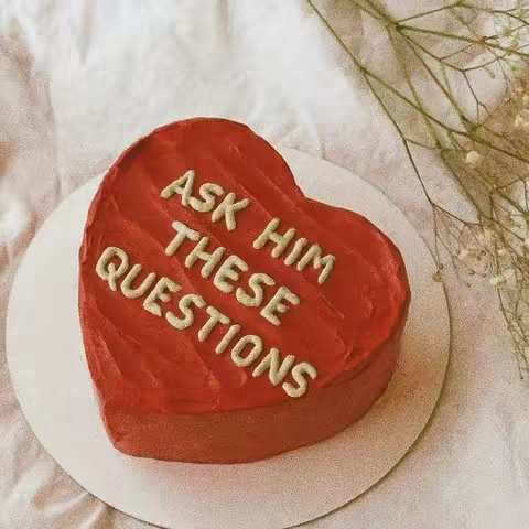 A heart-shaped red cake sits on a white, circular plate. The cake is decorated with white frosting, spelling out the words “ASK HIM THESE QUESTIONS”. The cake sits against a white background, with a few sprigs of delicate, dried flowers. The overall image has a warm, inviting, and slightly romantic feel.
